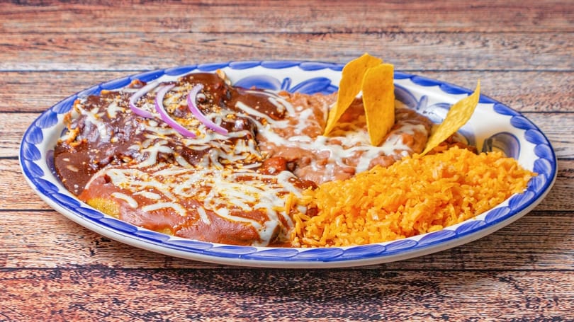 A plate of enchiladas with mole sauce, refried beans, rice, and tortilla chips.