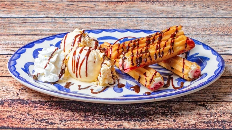Churros with chocolate drizzle, vanilla ice cream, and whipped cream on a blue and white plate.