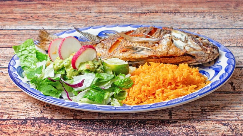 Fried whole fish with rice, salad, guacamole, radishes, and lime on a blue and white plate.