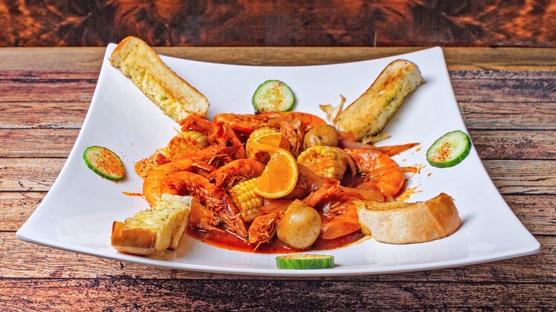 Shrimp boil with corn, potatoes, onions, and orange slices, served with garlic bread on a white plate.