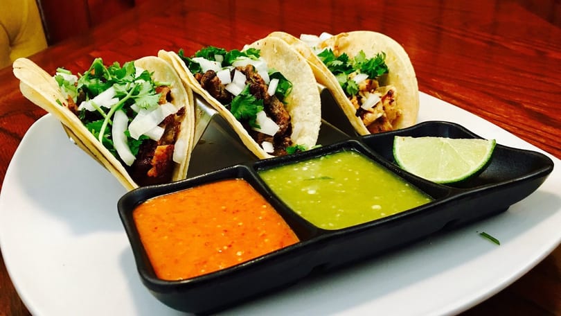 Four tacos with meat, onions, and cilantro, served with red and green salsa and a lime wedge.